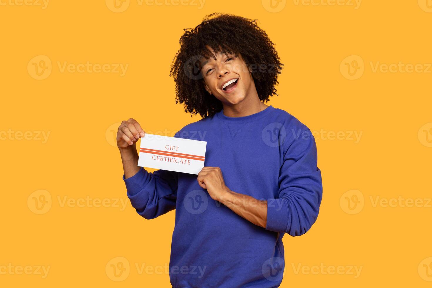 A cheerful young man, dressed in a blue sweatshirt, holds up a gift certificate while smiling broadly against a bright yellow backdrop. photo