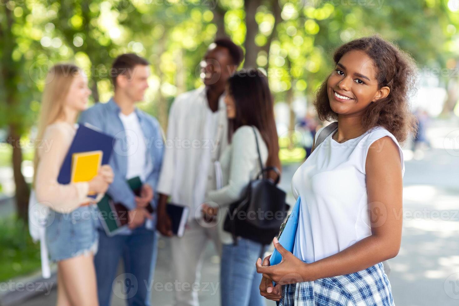 Happy african american girl student holding notepad and smiling at camera, spending time with friends in public park photo