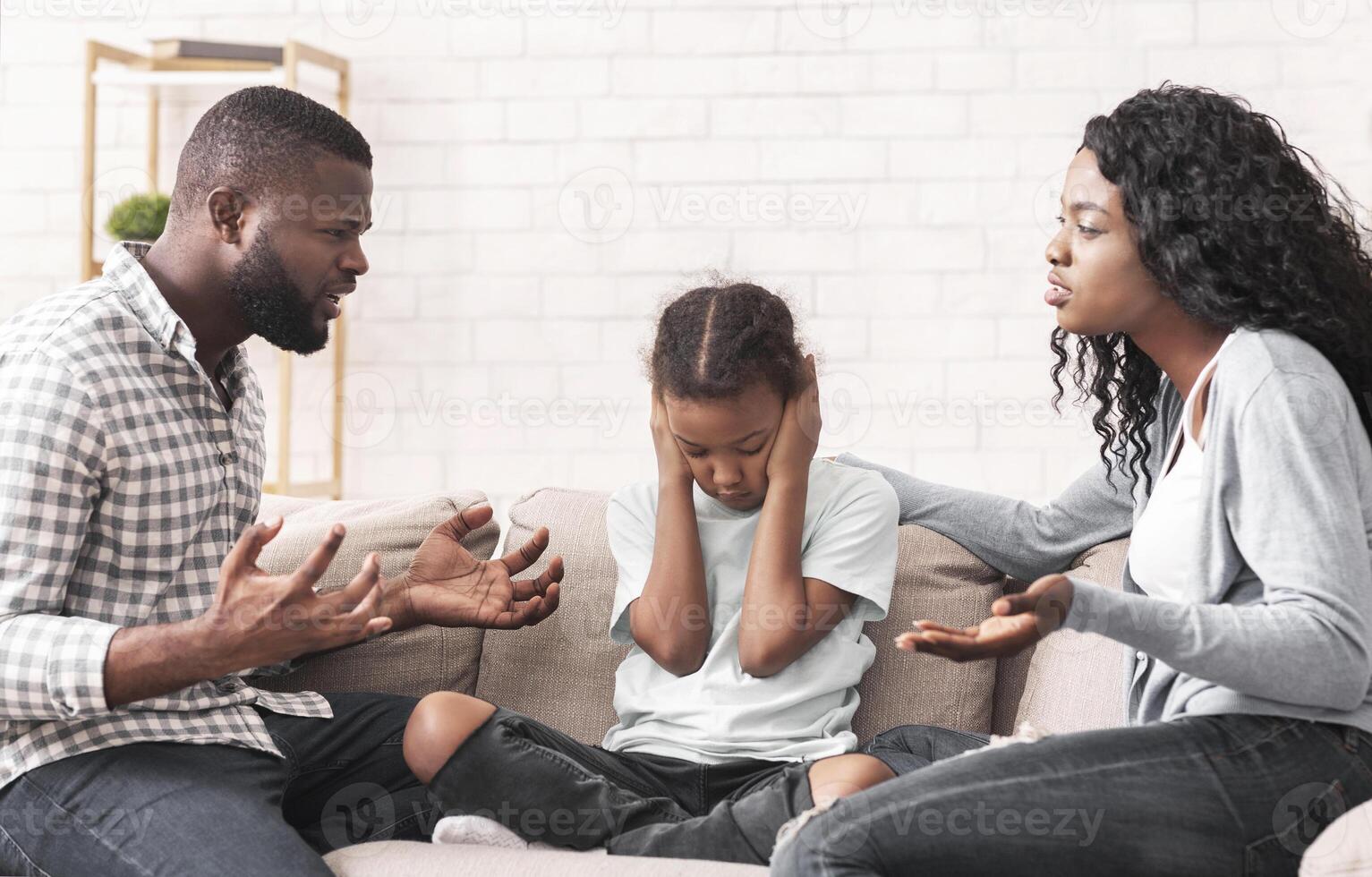 Depressed little black girl sitting between arguing parents, closing ears not to hear quarrels photo