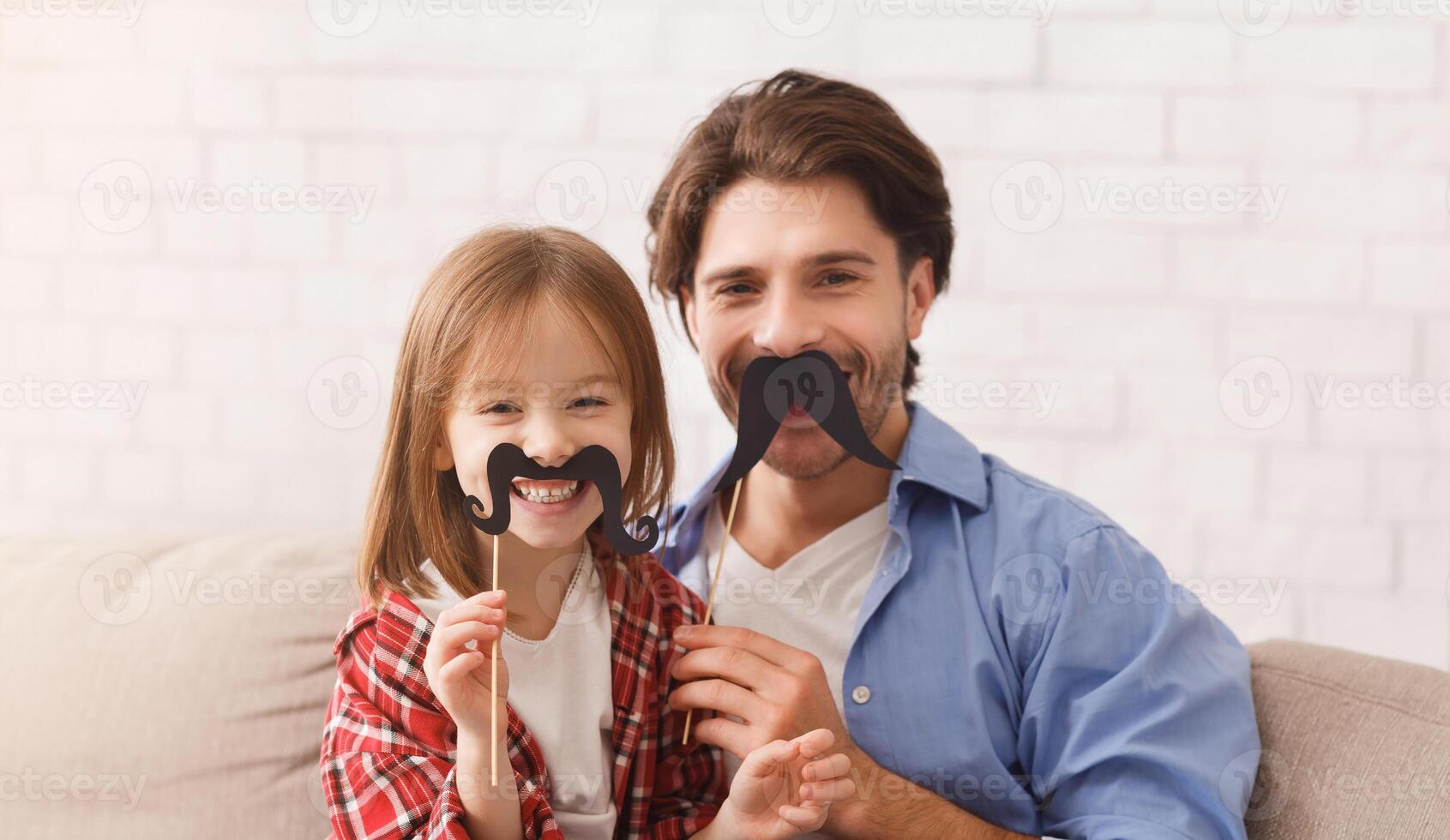 Beautiful funny little girl and daddy have mustaches on sticks, posing at home. Family holidays and togetherness concept, copy space photo