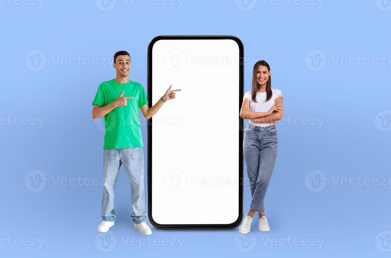 A smiling multiracial couple stand next to an oversized smartphone model, points at the empty screen, while the woman, in a white top and jeans, rests her elbow on the phone, indicating its features. photo