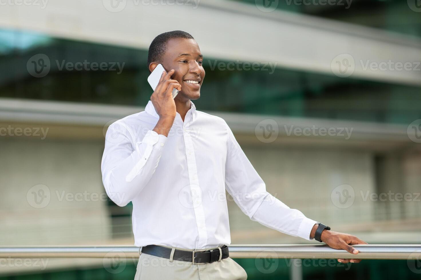 African American man in a white shirt and light-colored pants stands confidently, smiling while talking on his smartphone. The backdrop features a sleek glass building, copy space photo