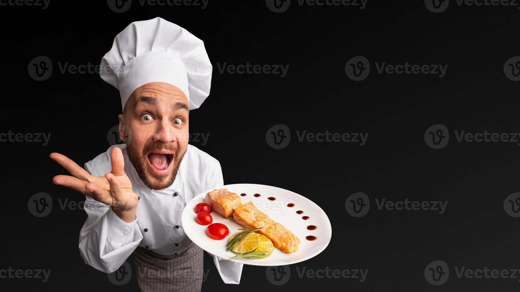 Funny Chef Man Showing Salmon Dish On Plate Posing In Studio On Black Background. High-Angle Shot photo