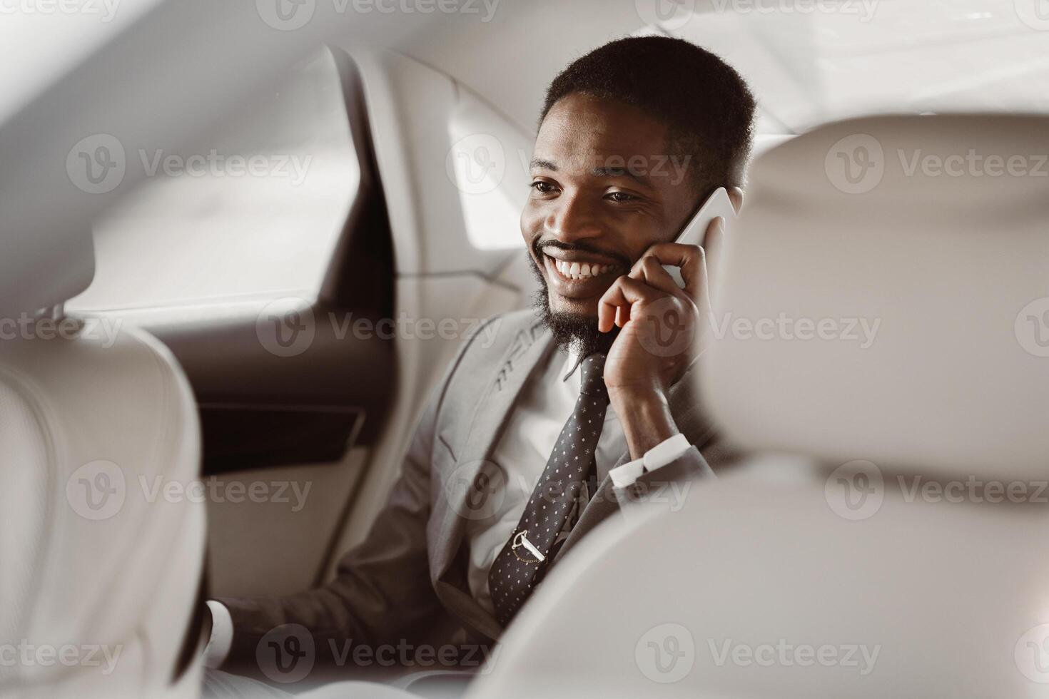 Successful African American businessman, wearing a suit and tie, sits in the backseat of a car and talks on a phone while smiling. photo