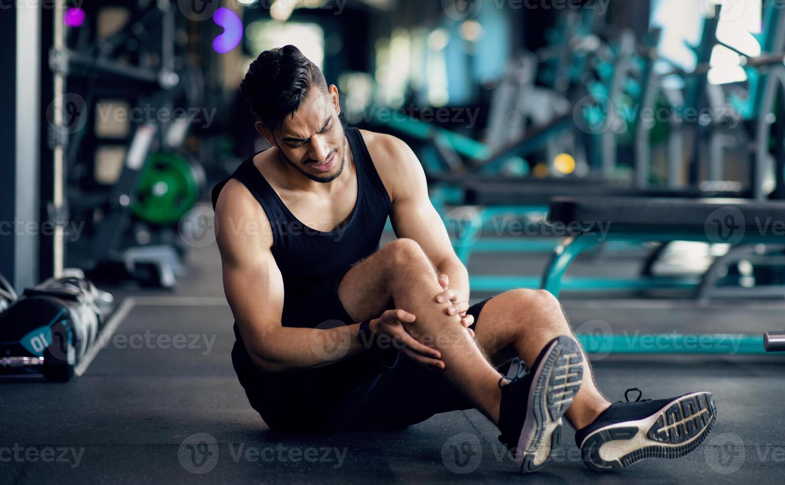 Sport Injury. Upset Arab Male Athlete Suffering From Knee Trauma While Training At Gym, Middle Eastern Man Sitting On Floor And Massaging Aching Area, Sporty Guy Frowning With Pain, Copy Space photo