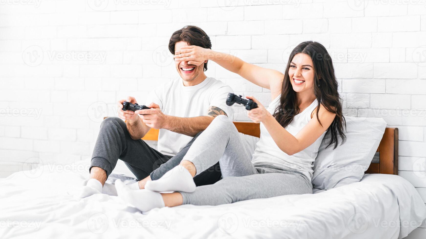 A happy couple is playing games in bed at home. The woman is laughing as she holds her controller up while her boyfriend is trying to focus on the game. photo