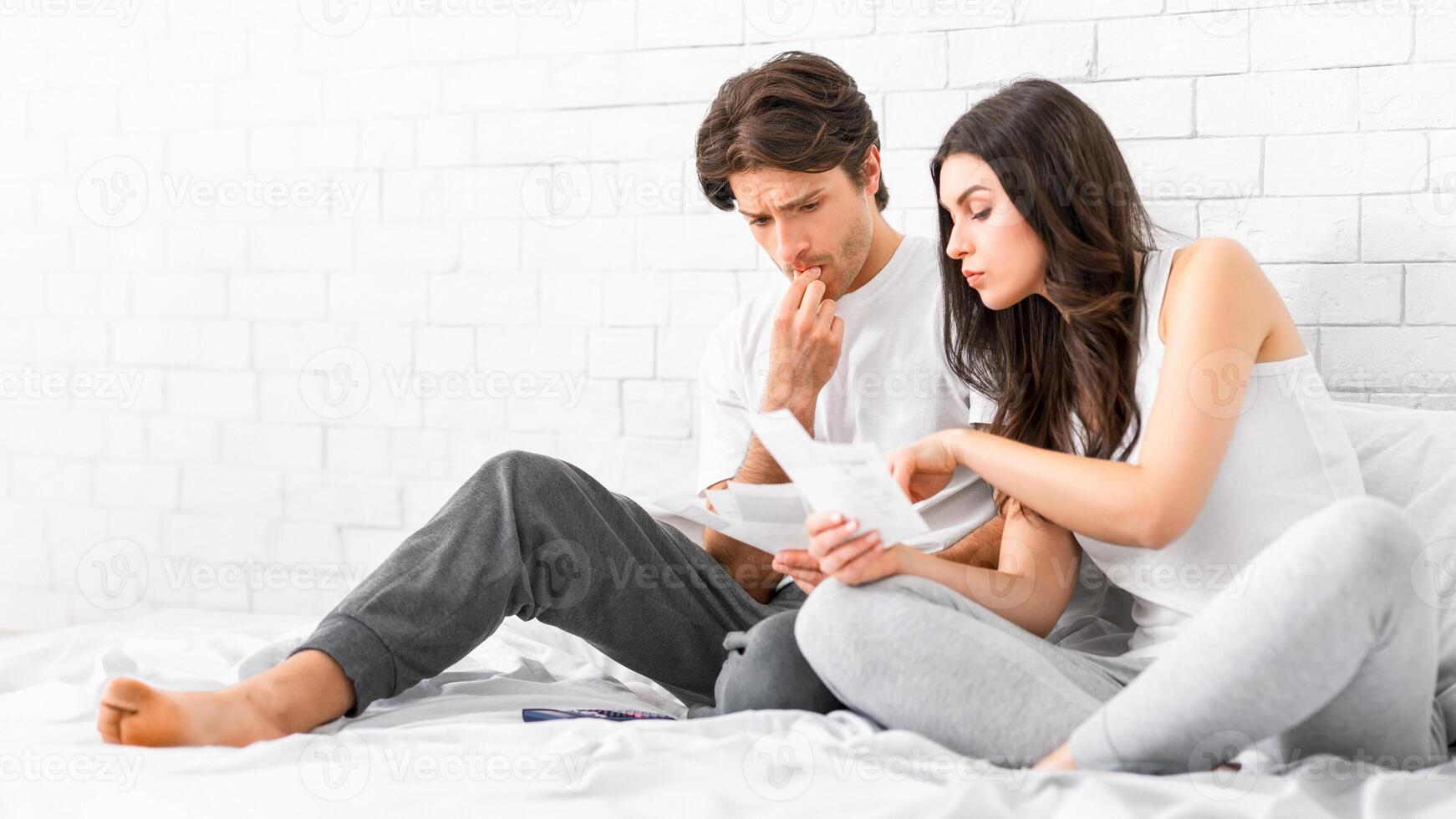 A young couple sits together on a white bed, looking at bills and documents. The man, wearing a white shirt and grey pants, is biting his fingernail and appears stressed. photo