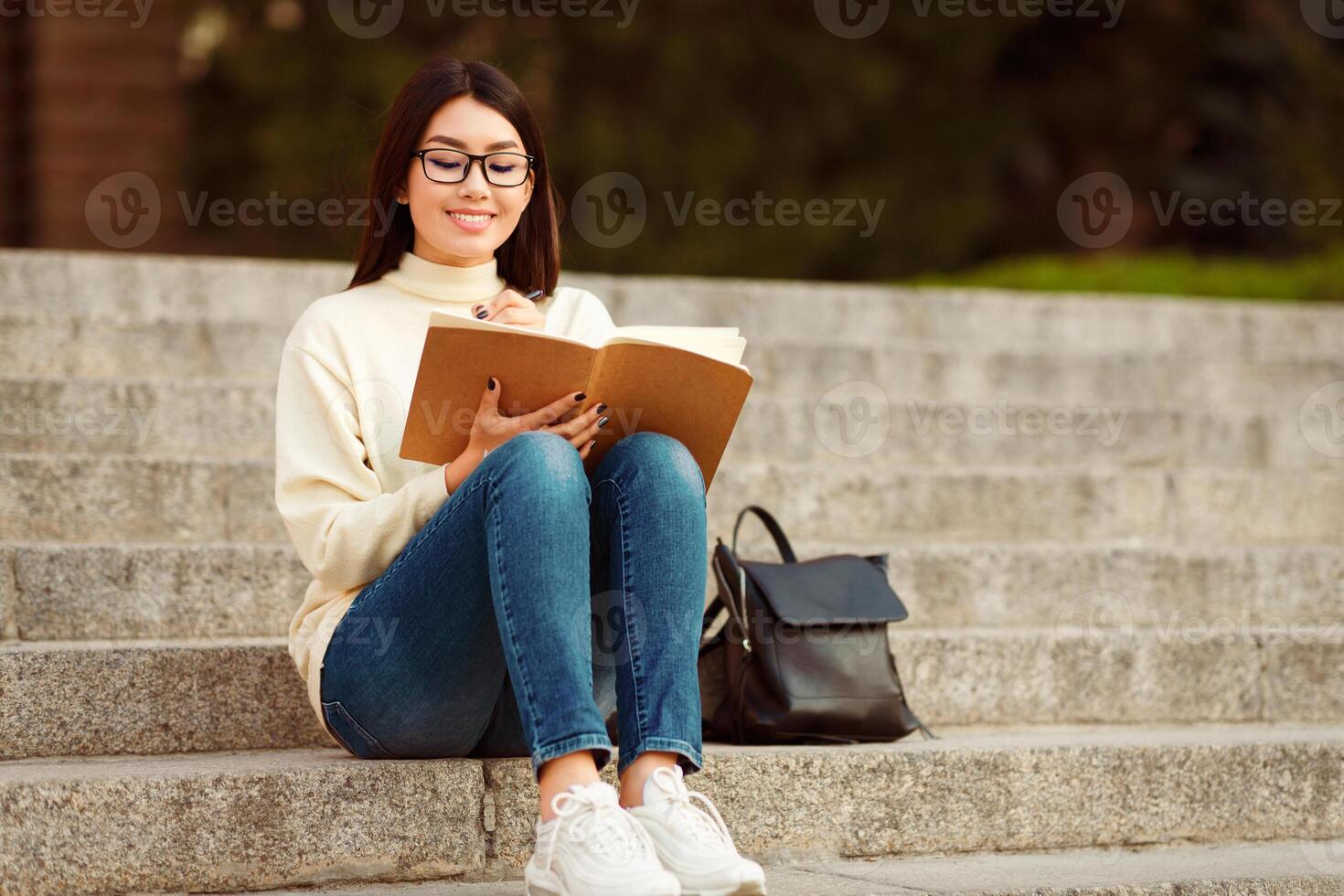 Asian woman student is seated on a set of steps, engrossed in reading a book. She is holding the book in her hands and appears focused on the pages. photo