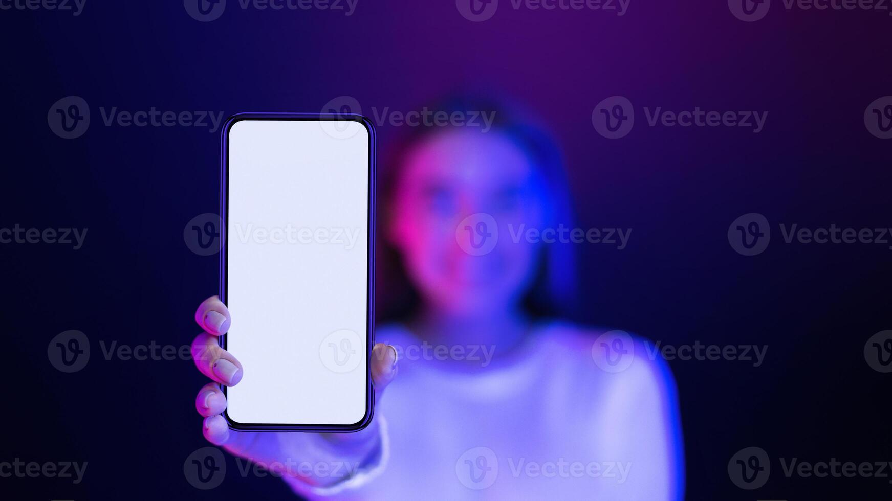 A woman is standing in front of a purple background, holding up a phone with a blank screen. She appears to be showcasing the device, mockup, copy space photo