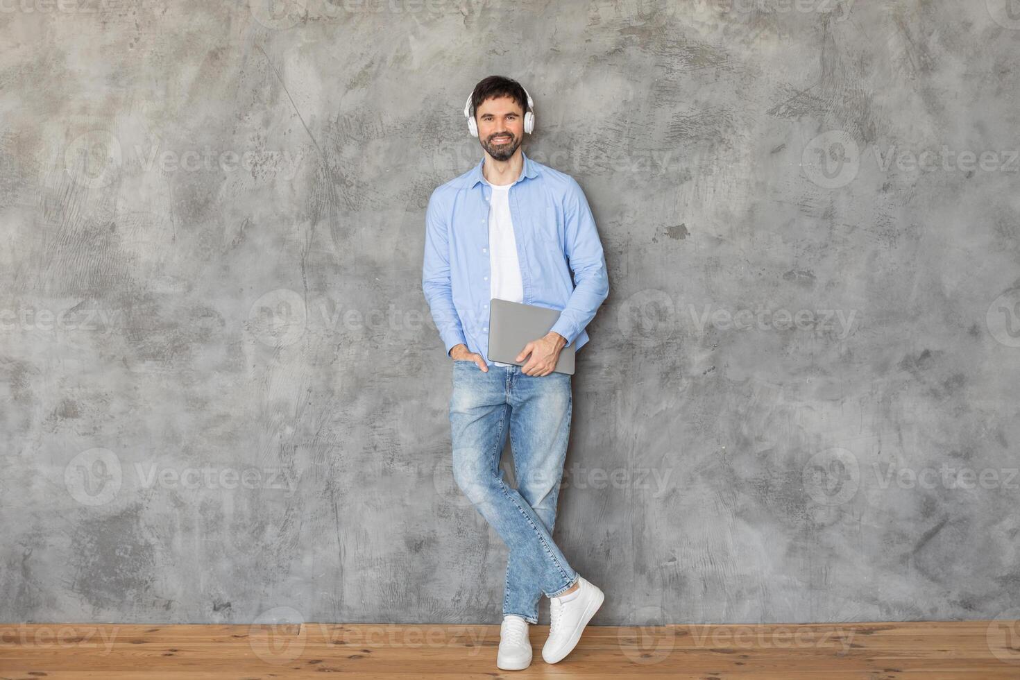 Man standing in front of a concrete wall, leaning against it with one hand in his pocket. He is smiling at the camera and wearing headphones over his ears. He is holding a laptop in his other hand photo