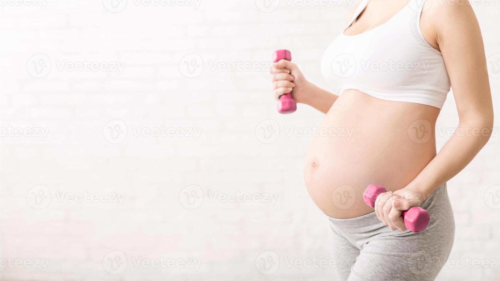 A pregnant woman is shown holding two pink dumbbells in a fitness setting. She appears to be engaged in weightlifting exercises as part of her prenatal workout routine, cropped, copy space photo