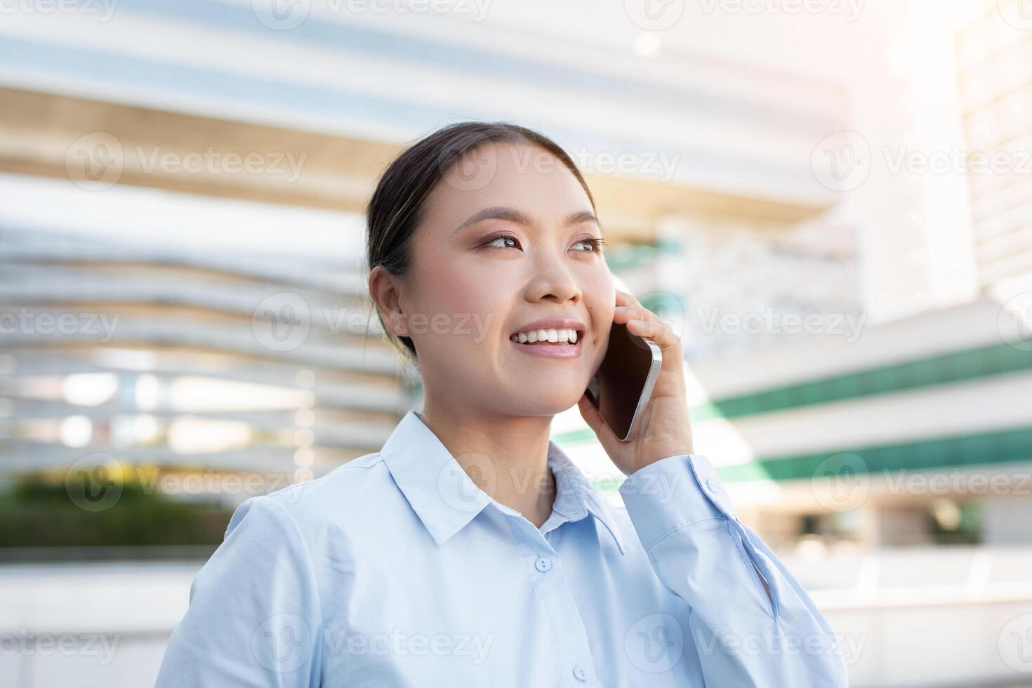 A young Asian lady professional with a friendly smile is engaged in a lively phone conversation outside. The modern architecture in the background suggests an urban environment photo
