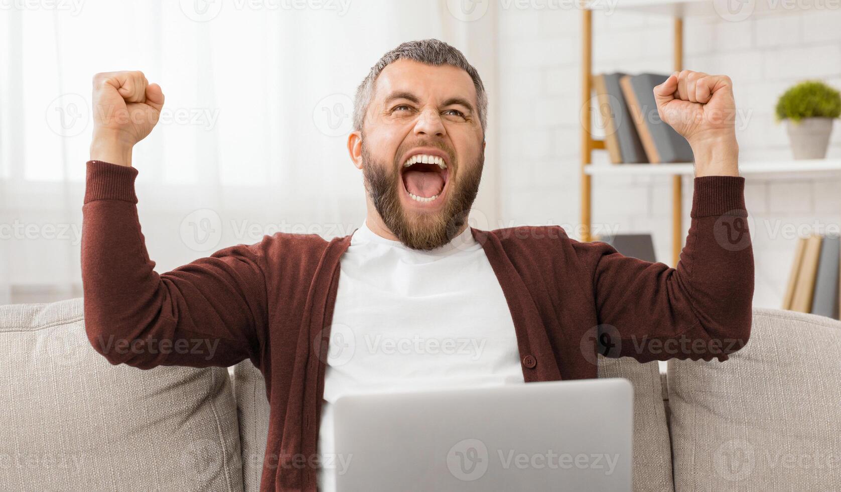 A man is seen sitting on a couch with laptop and his arms raised in the air. He appears to be energetic and possibly celebrating something. The background is a simple living room setting. photo