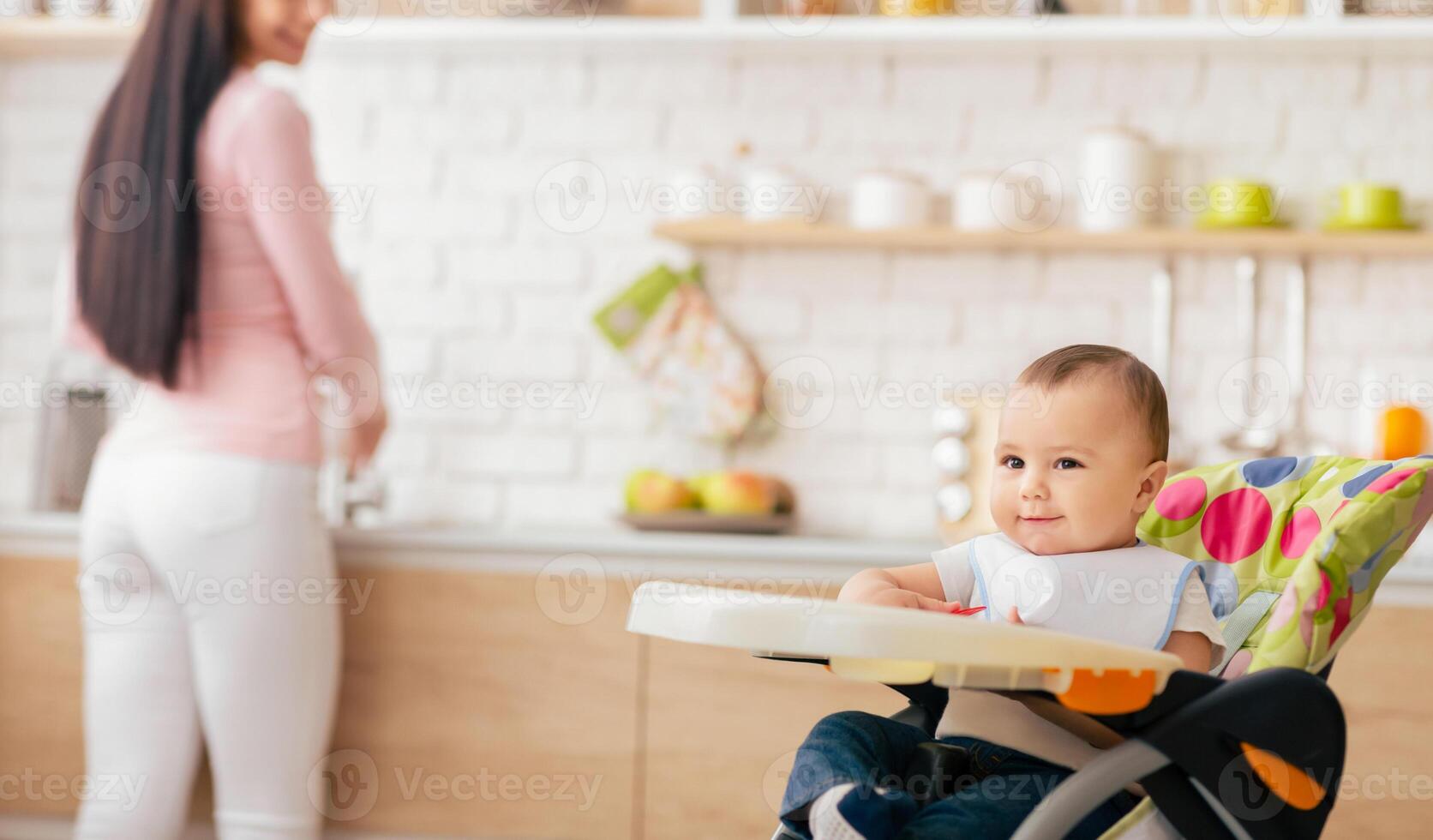 A smiling baby sits comfortably in a colorful high chair in a modern kitchen while the mother is busy in the background preparing food. photo