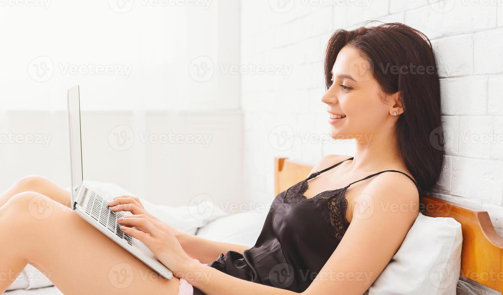 A woman is sitting on a bed while using a laptop computer. She appears focused on the screen, typing and clicking. The room is simple and uncluttered, with soft lighting from a nearby window. photo