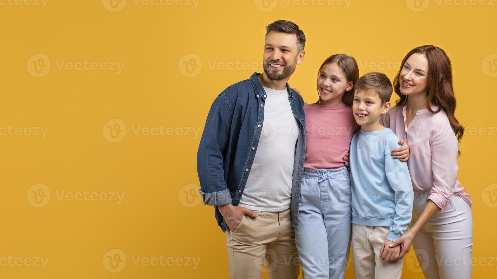 A happy family, with two parents and two children, embrace each other against a vibrant yellow backdrop. They appear cheerful and content, looking at copy space photo