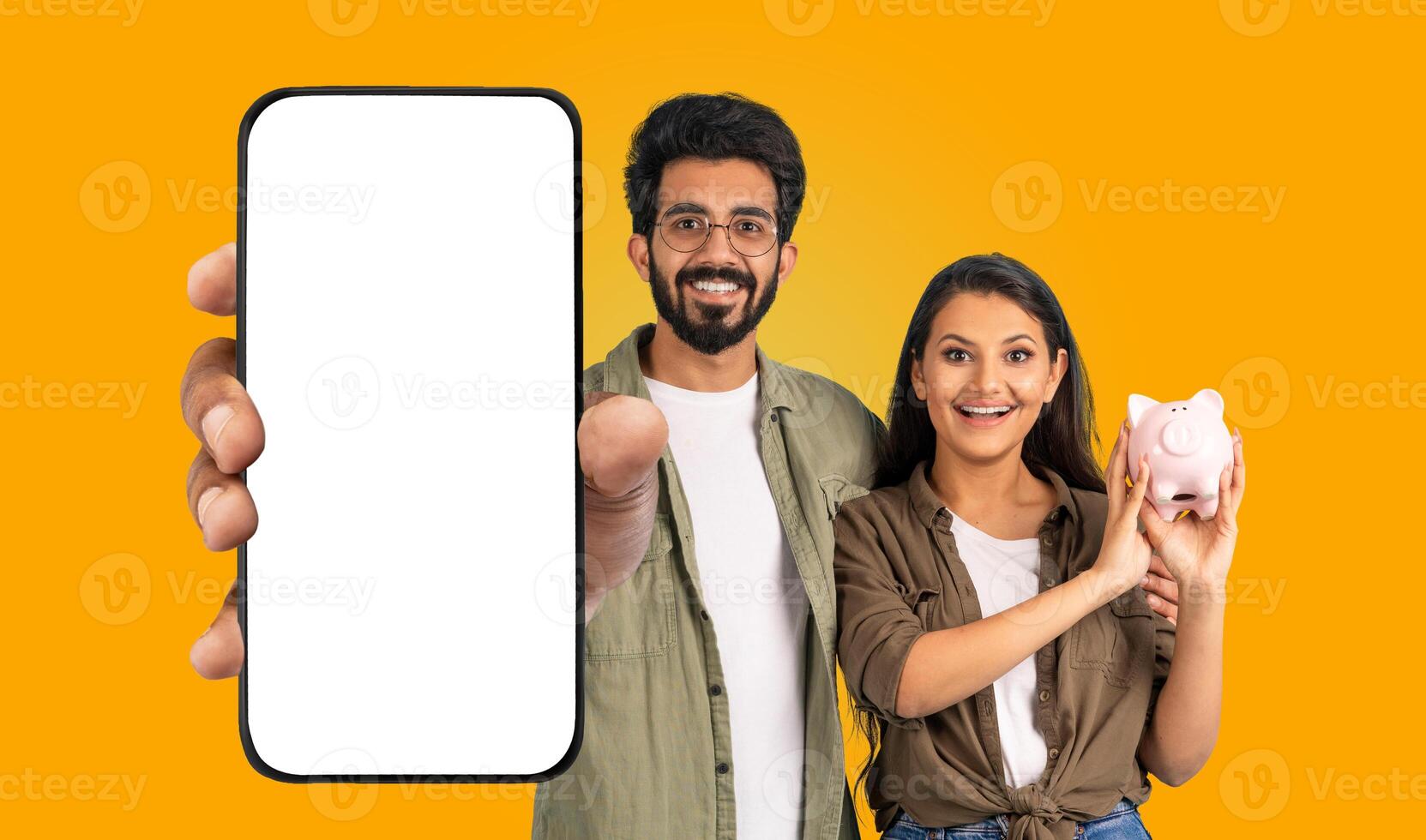 A young Hindu man and woman smile and look at the camera. The man holds a smartphone with a blank white screen and points his finger at the screen. The woman holds a pink piggy bank in her hand. photo