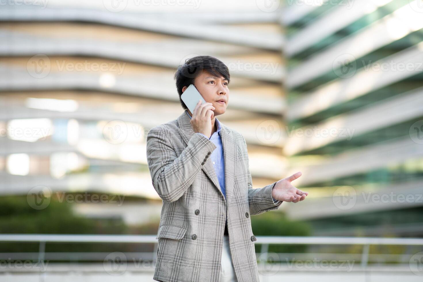 Asian young man wearing a plaid jacket is standing outside of a modern building, talking on his cell phone while gesturing with his other hand. photo
