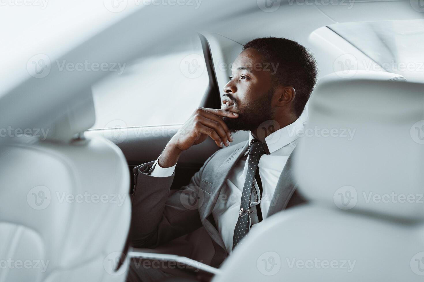 A black man in a grey suit with a white shirt and a dark tie sits in the backseat of a car looking out the window, deep in thought. His hand rests on his chin as he ponders. photo