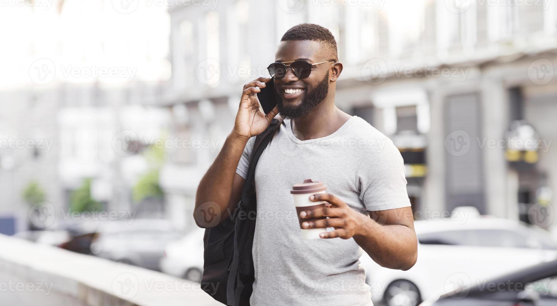 A man in sunglasses smiles while talking on a cell phone and holding a coffee cup in his hand. He is walking down a city street, and cars can be seen in the background. photo