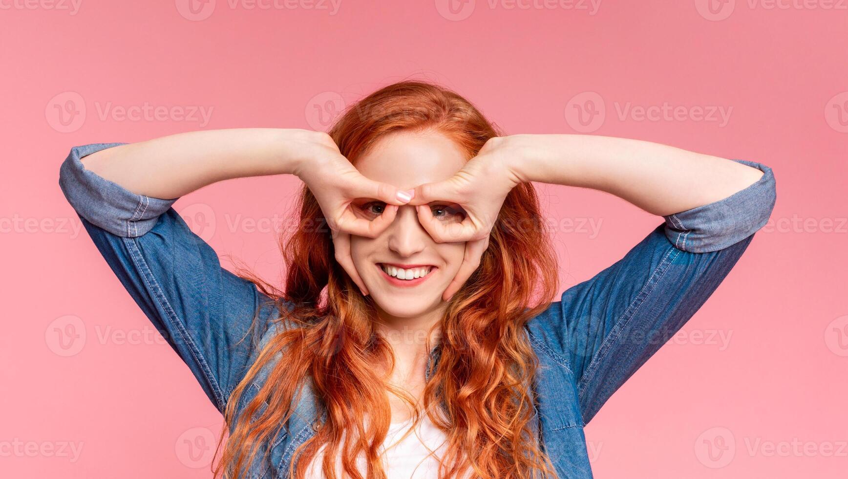 A cheerful redheaded woman is standing against a solid pink backdrop, forming specs with her hands around her eyes photo