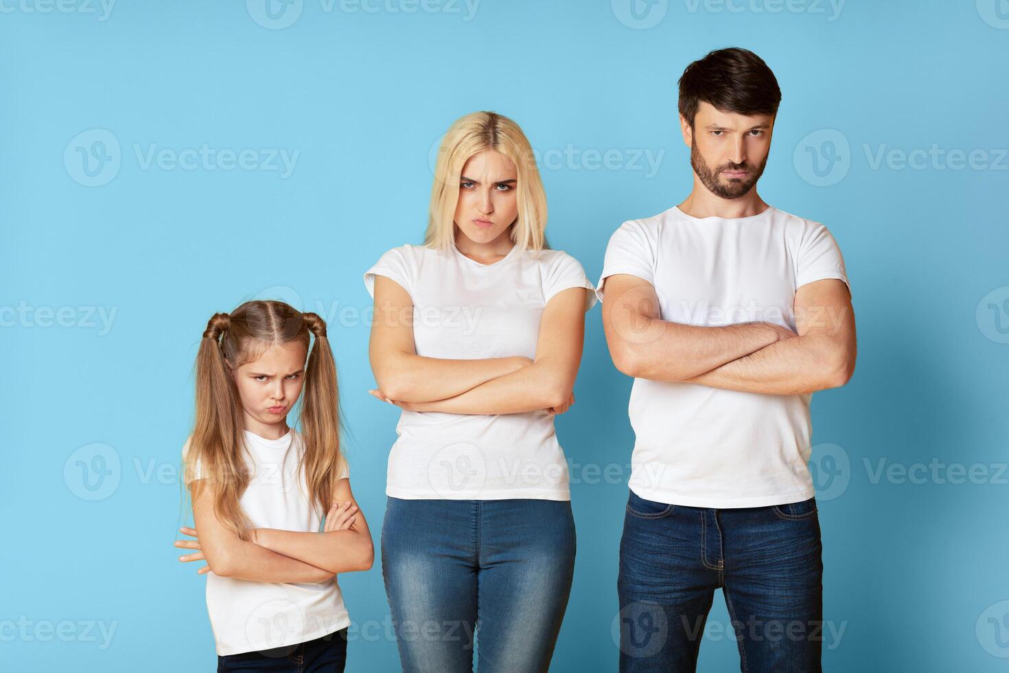 A family of three stands in front of a light blue background with their arms crossed. The young girl has a frustrated expression on her face. In the middle is the mother appears to be annoyed. photo
