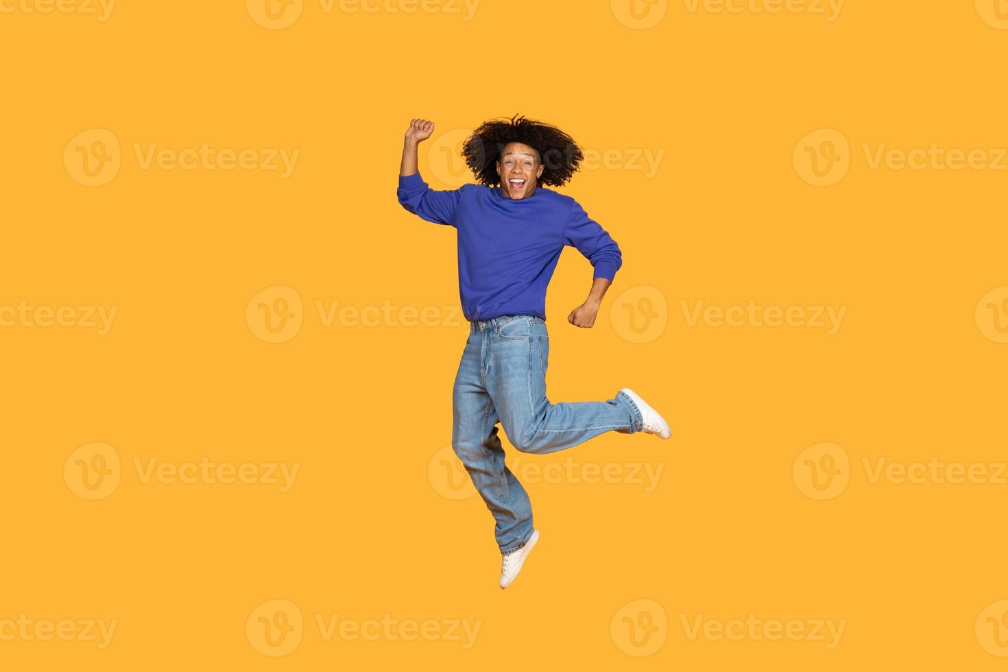 A young man with curly hair, wearing a blue sweater and jeans, jumps in the air with a big smile on his face, against a bright yellow background. He appears to be in a moment of pure joy photo