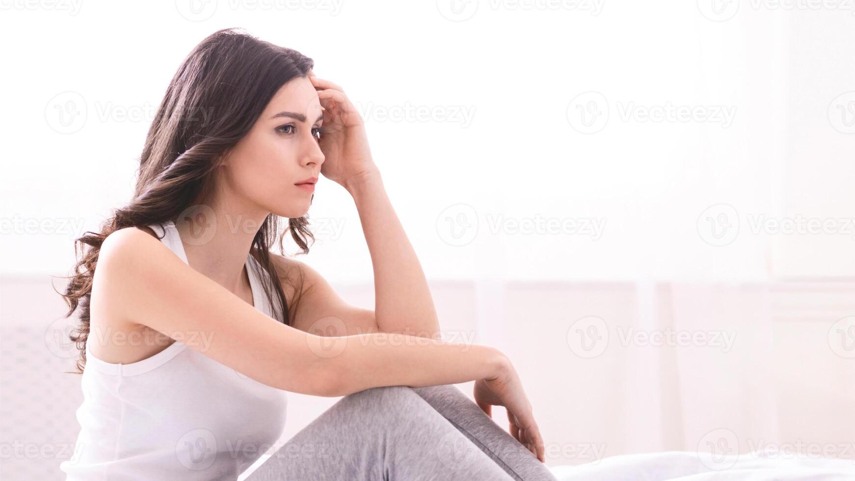 A woman with long brown hair sits on a bed, her hand resting on her forehead. Her expression is one of worry or sadness. She is wearing a white tank top and grey pants. photo