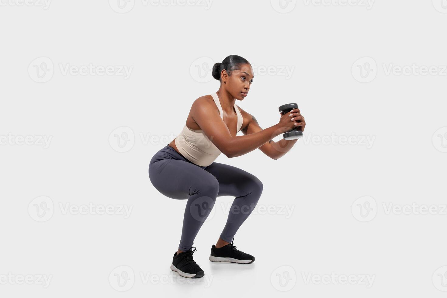 African American woman is doing a squat exercise while holding a dumbbell in her hand. She is lowering her body down by bending her knees and hips, then pushing back up to the starting position photo