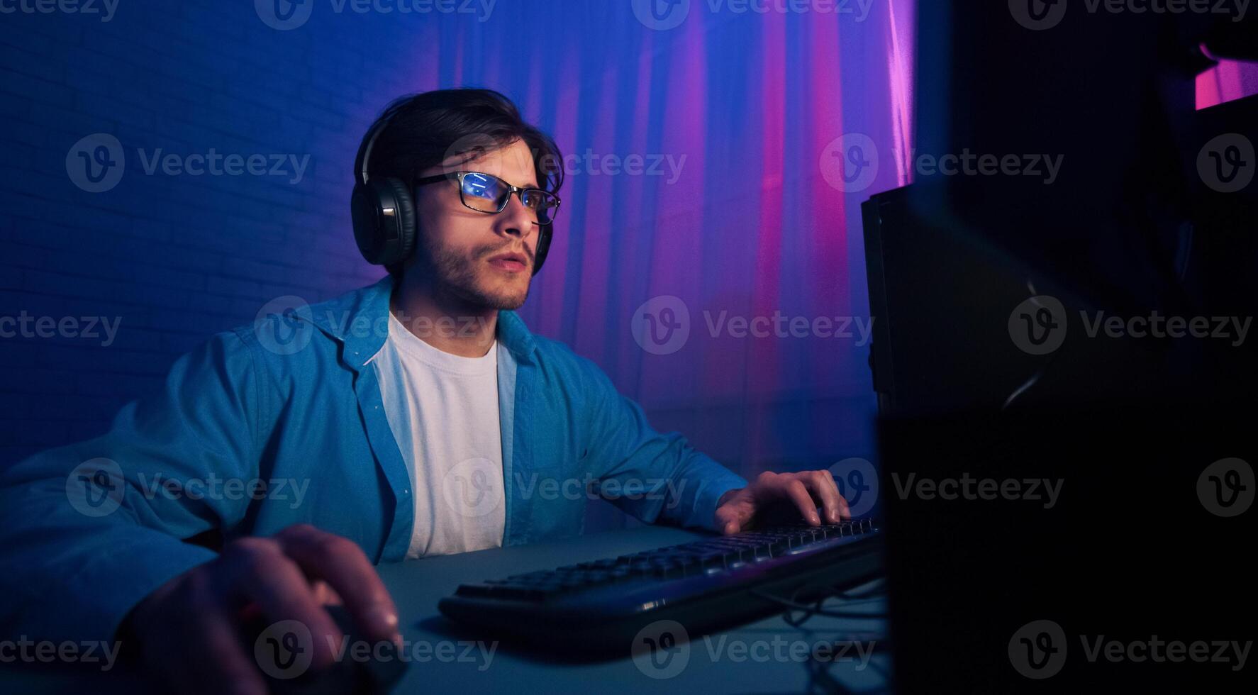 A man wearing a blue shirt and glasses is focused on his computer screen. He is using a keyboard and mouse, and has headphones on. The room is lit with blue and pink neon lights. photo