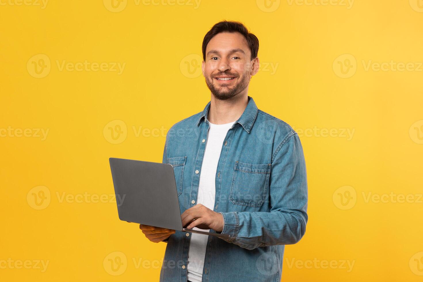 A man wearing a blue shirt is shown holding a laptop with both hands. The man appears focused on the screen of the laptop, possibly working or browsing photo