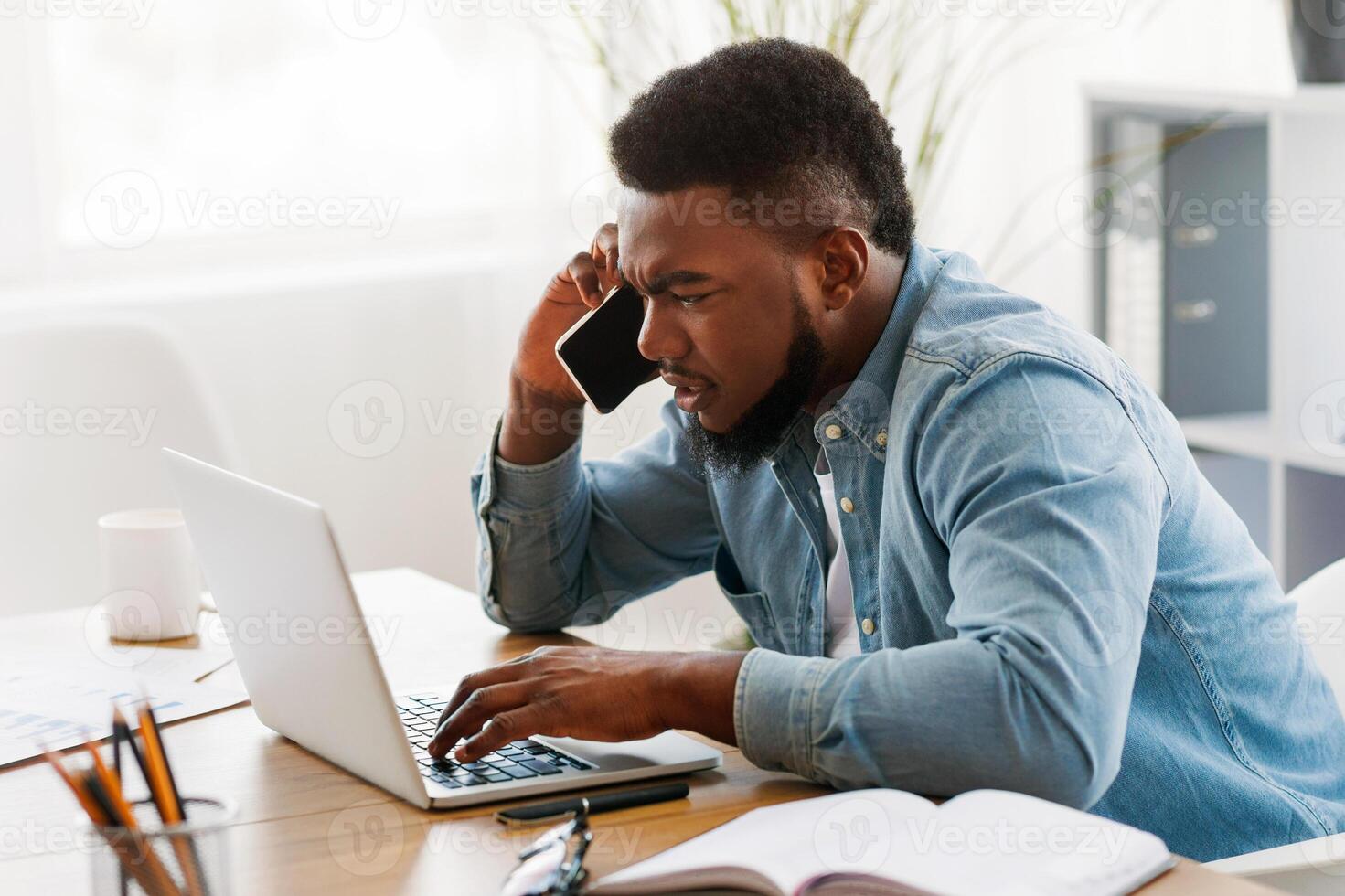 Worried black businessman talking on cellphone and looking at laptop screen, trying to solve problems online, side view with copy space photo