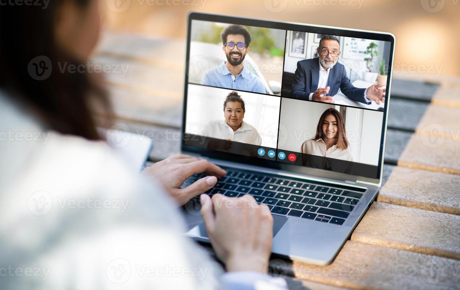 Woman is participating in a virtual meeting on a laptop placed on an outdoor table. Four colleagues are visible on the screen, each engaging in conversation from different locations photo