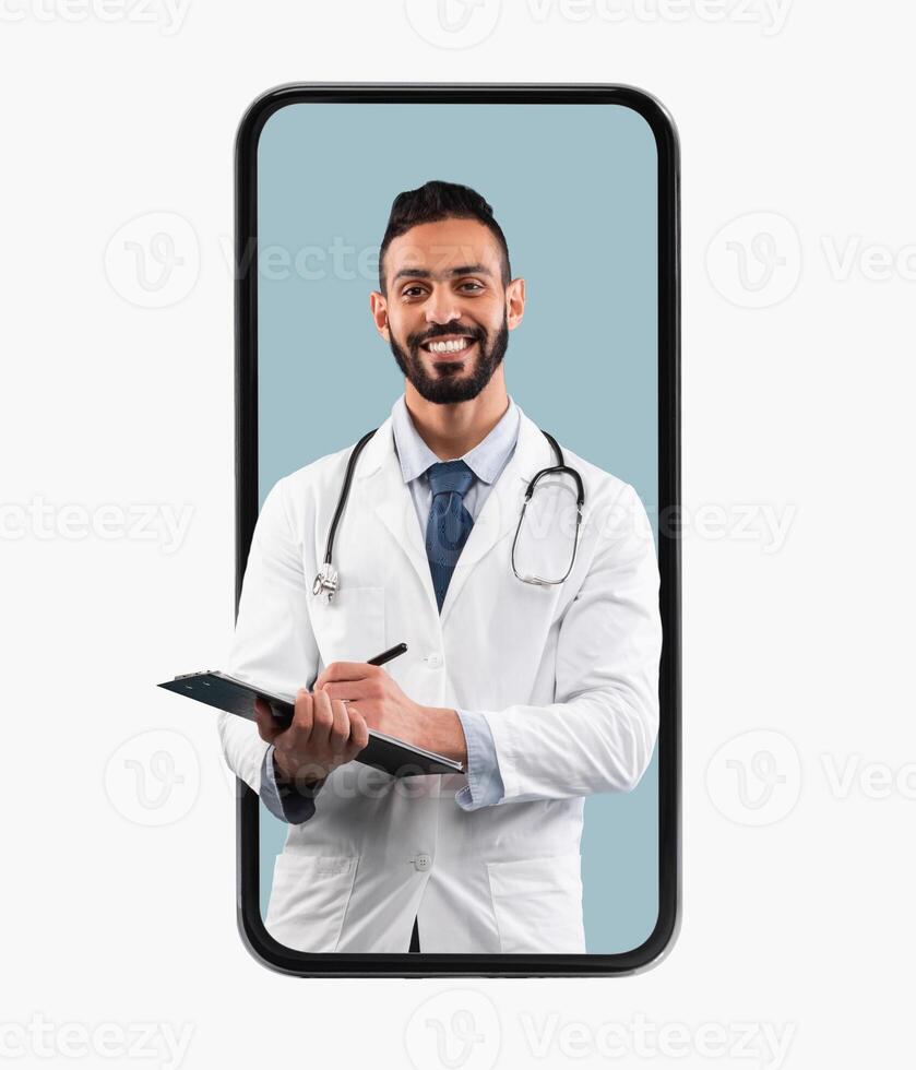 Medical Services. Young Happy Male Middle Eastern Doctor In White Uniform With Stethoscope And Clipboard Writing Prescription To Patient And Smiling At Camera, Looking Coming Out Big Cell Phone Screen photo