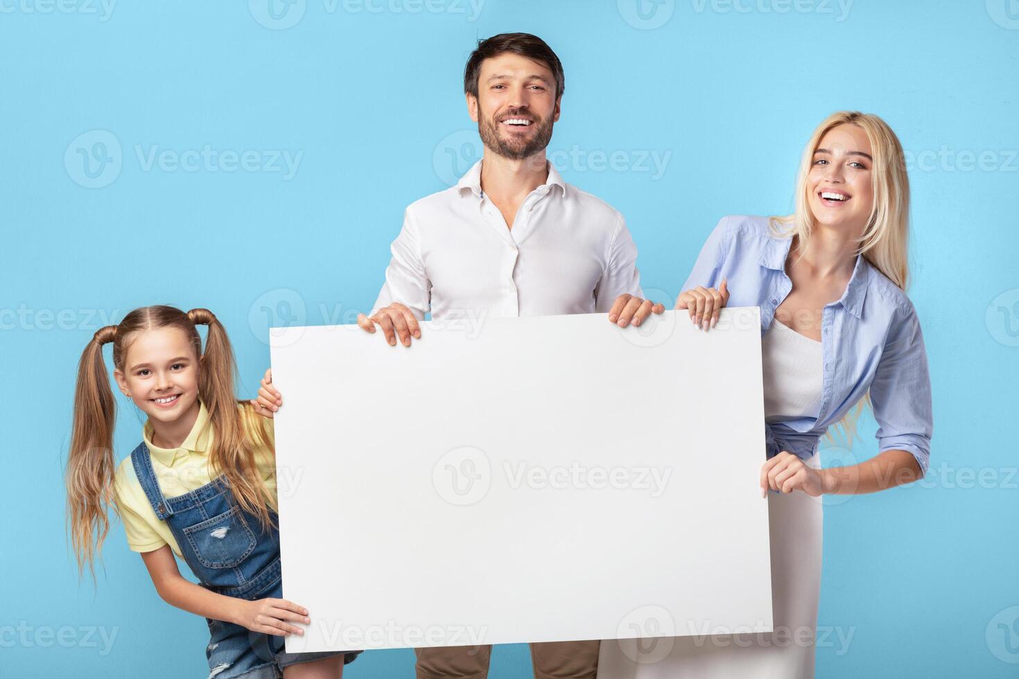 A happy family of three is standing in front of a blue background, holding a large, blank sign. They are all looking at the camera. photo