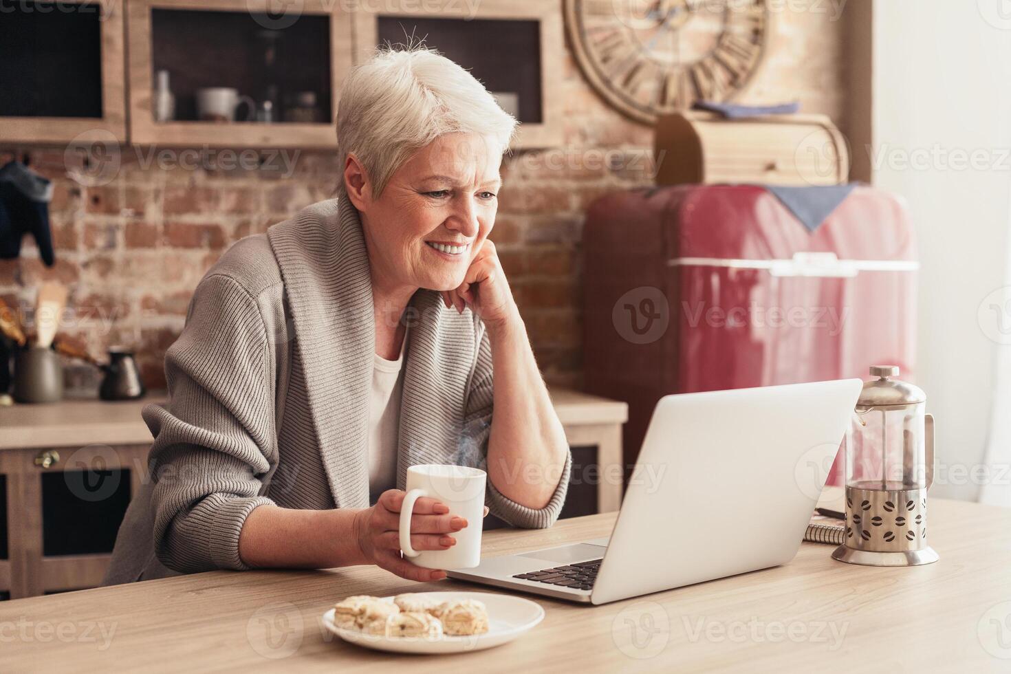 A senior woman with short, gray hair is sitting at a kitchen table, smiling as she looks at her laptop. She is holding a white mug of coffee and there is a plate of pastries beside her computer photo