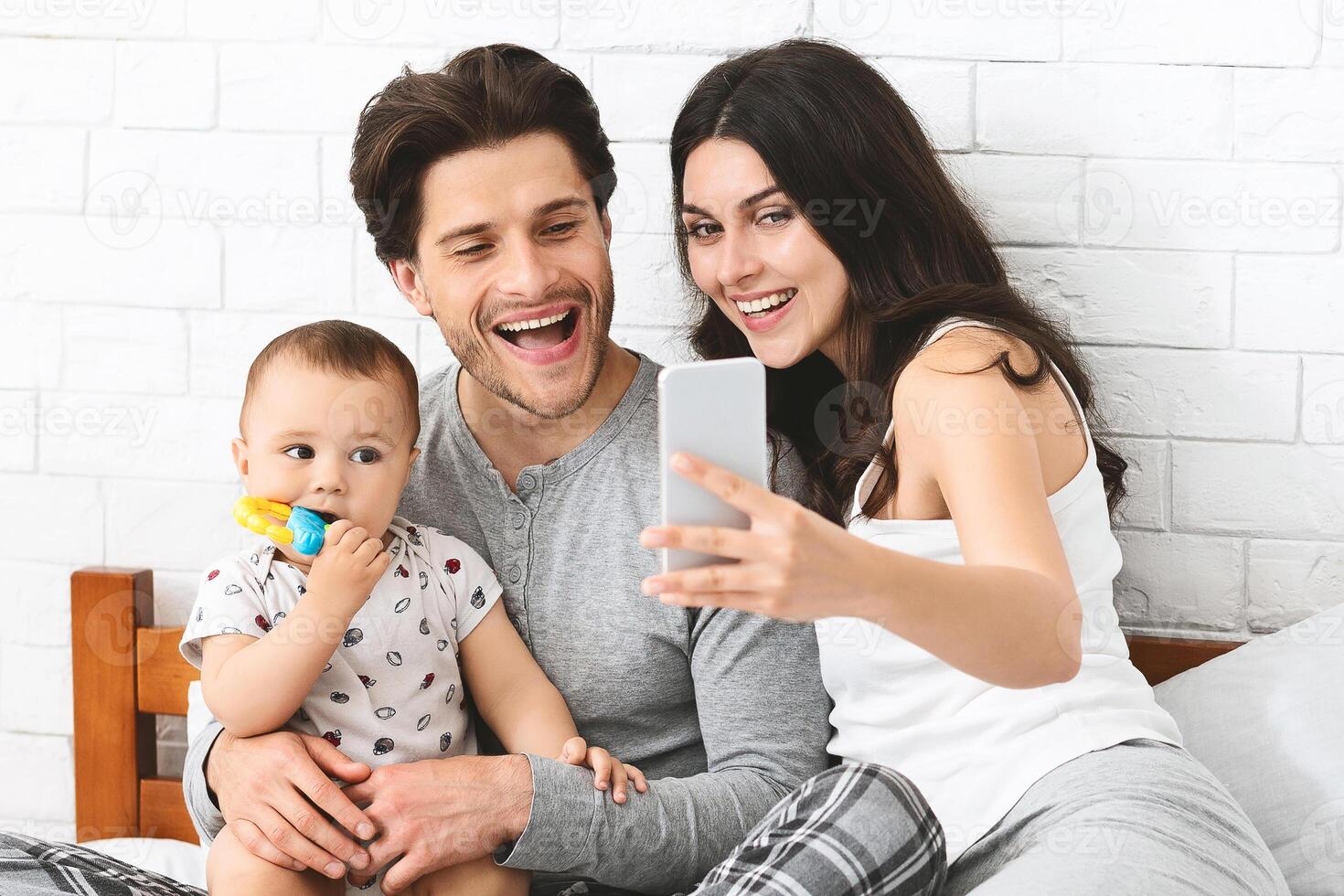 This image shows a happy family taking a selfie in their bedroom. A young man holds his baby son in his lap while his wife takes the picture on a smartphone. photo