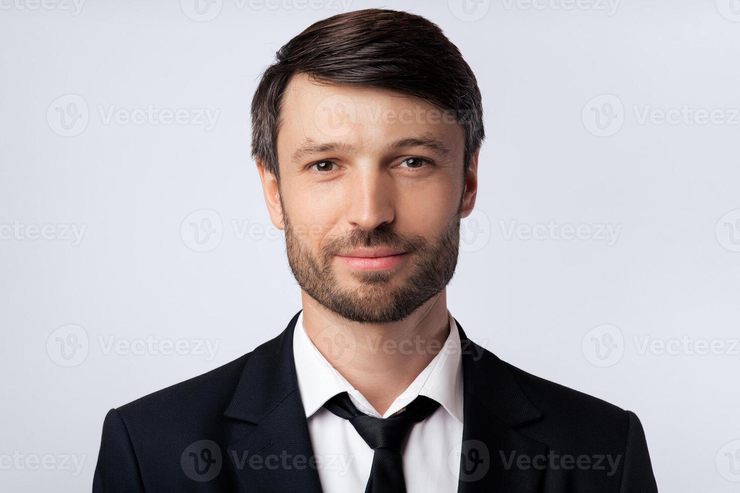 A close-up portrait of a man wearing a black suit, white shirt, and a black tie, smiling subtly. The background is a solid white. photo
