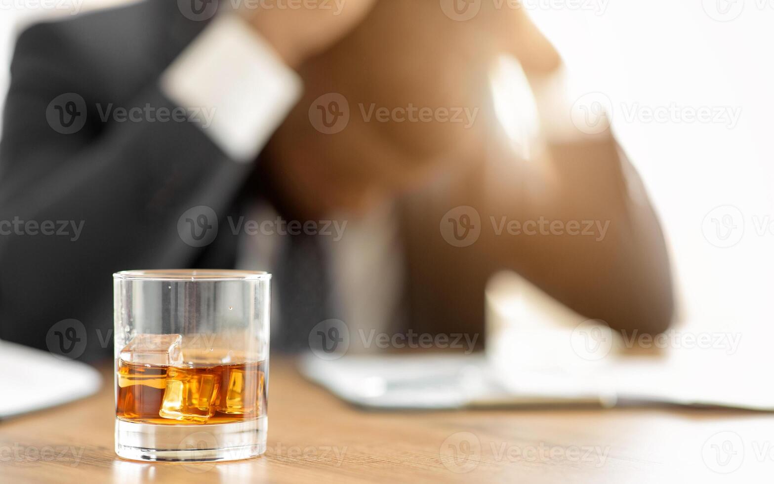 A glass of whiskey sits on a desk with ice cubes. A person is out of focus in the background. photo