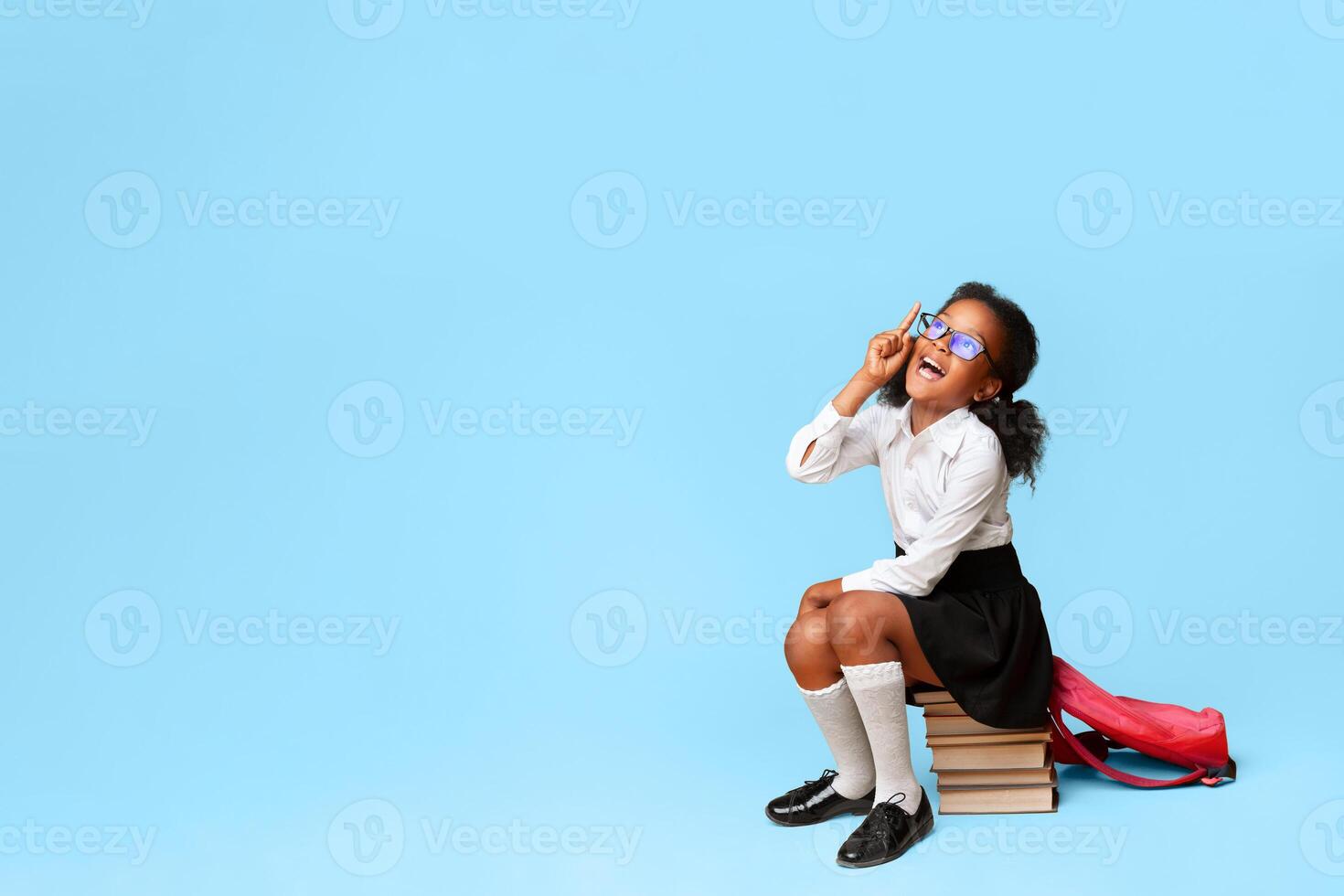 African Schoolgirl Having Idea Pointing Finger Up Sitting On Books Over Yellow Background. Copy Space, Studio Shot photo