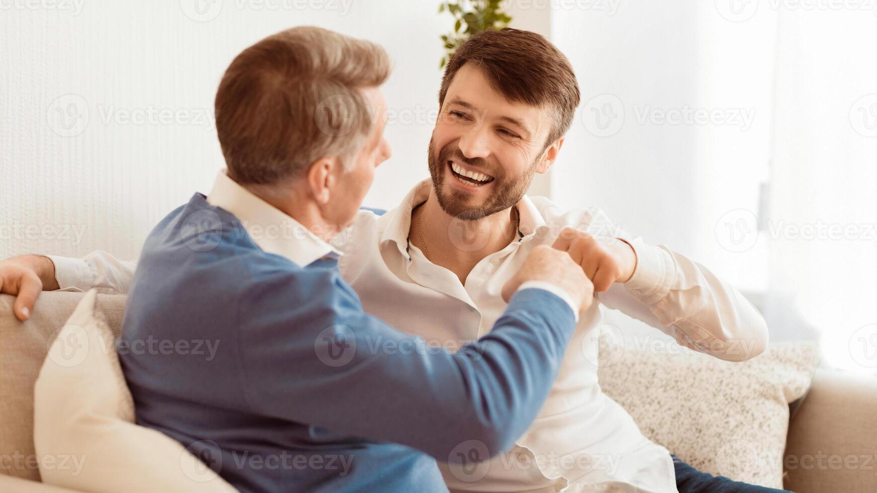 Mature Man And Senior Father Bumping Fists Sitting On Sofa At Home. Spending Time With Elderly Parent. Panorama, Selective Focus photo