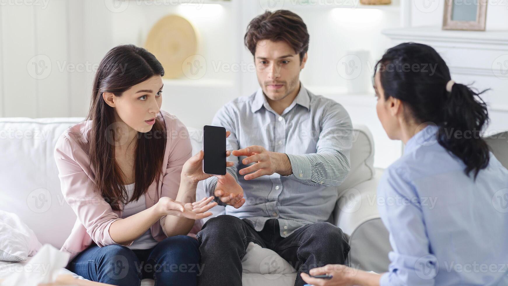 A young couple sits on a white sofa in a cozy living room, holding a smartphone and gesturing while discussing with a consultant. Shelves and a white wall are in the background. photo