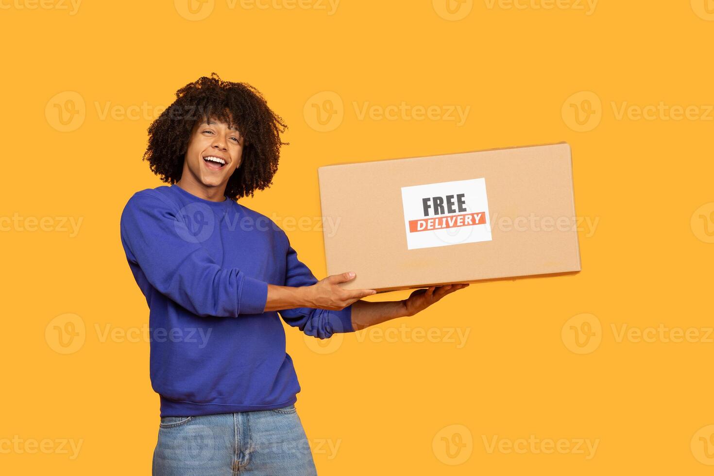 A young man with curly hair smiles broadly while holding a large cardboard box labeled with a free delivery sign. The bright yellow background, the excitement of receiving a package. photo