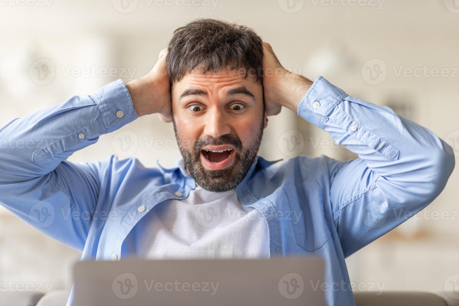 A man sits in front of a laptop, staring at the screen with shock and surprise. He holds his head in his hands with a wide-eyed expression and an open mouth, closeup photo