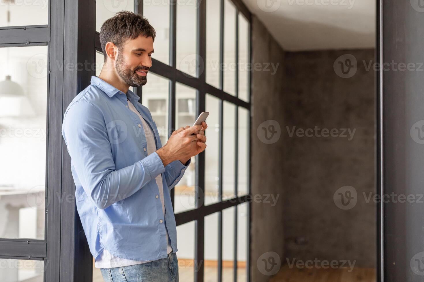 A man wearing a blue button-up shirt and jeans stands near a large glass door in a modern apartment. He is looking down at his smartphone and smiling, copy space photo