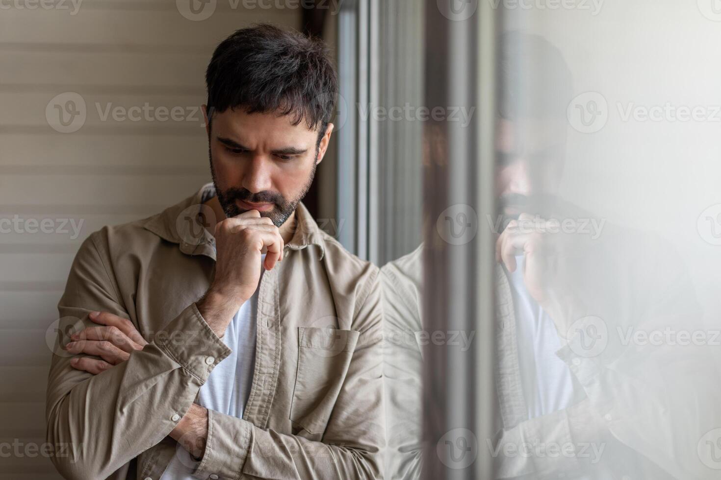 A man stands in front of a window, deep in thought, with his hand resting on his chin. The natural light illuminates his profile as he gazes outside, deep in contemplation. photo