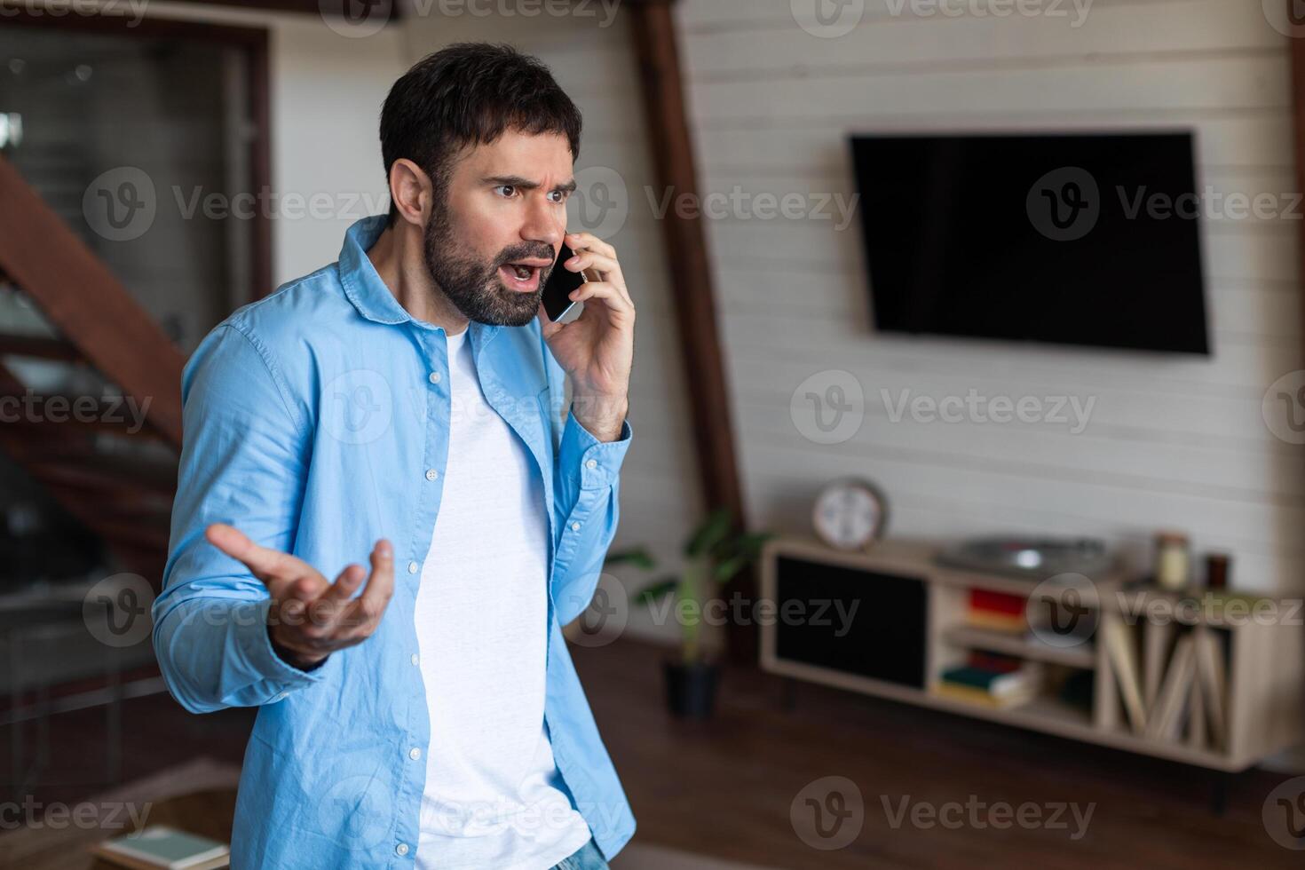 A man stands in a well-furnished living room, expressing concern or surprise as he talks on a mobile phone. His body language suggests an intense or possibly stressful conversation photo