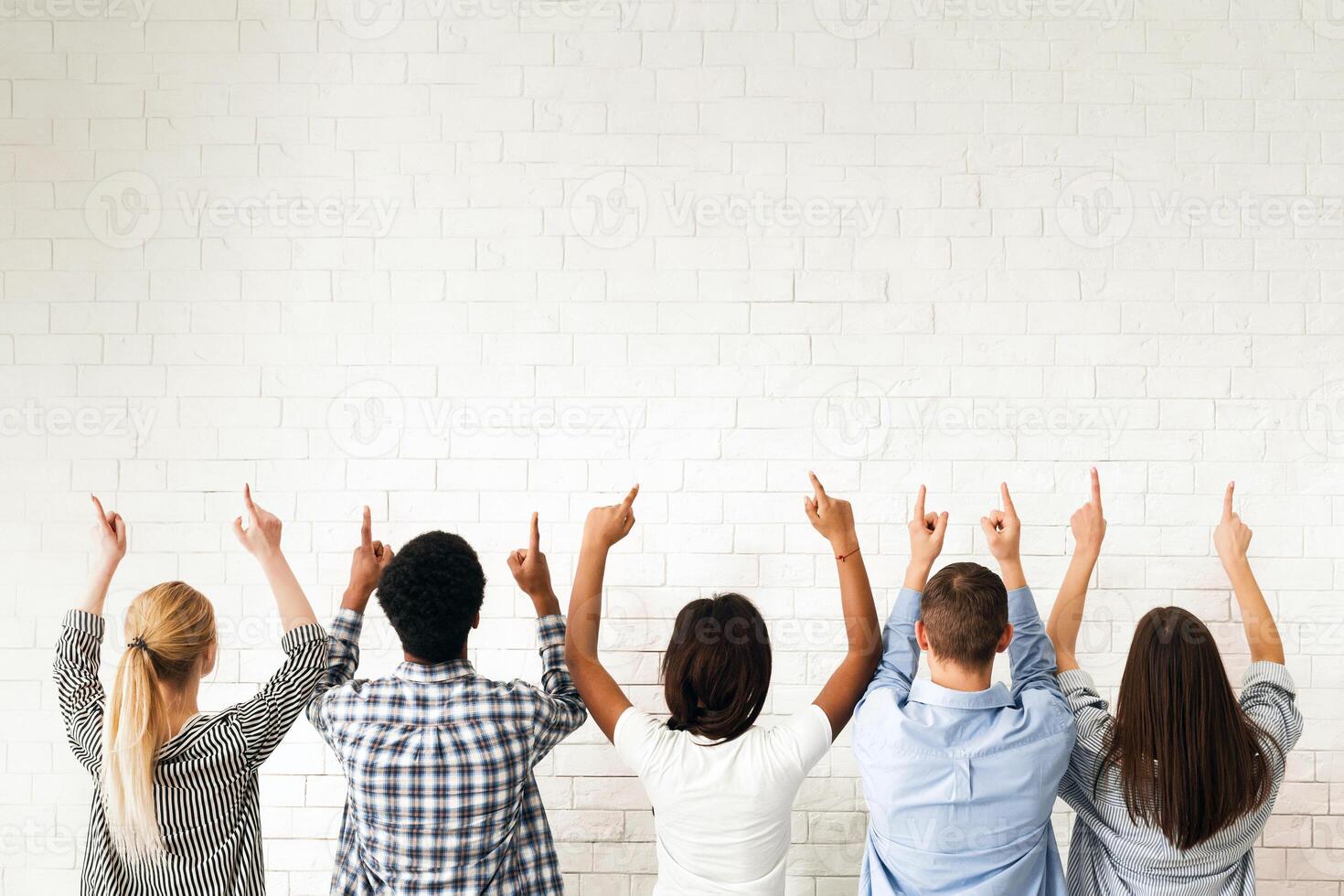 A group of friends stands in front of a blank white wall, all pointing upwards. Their gestures suggest they are indicating or drawing attention to something above them, back view photo