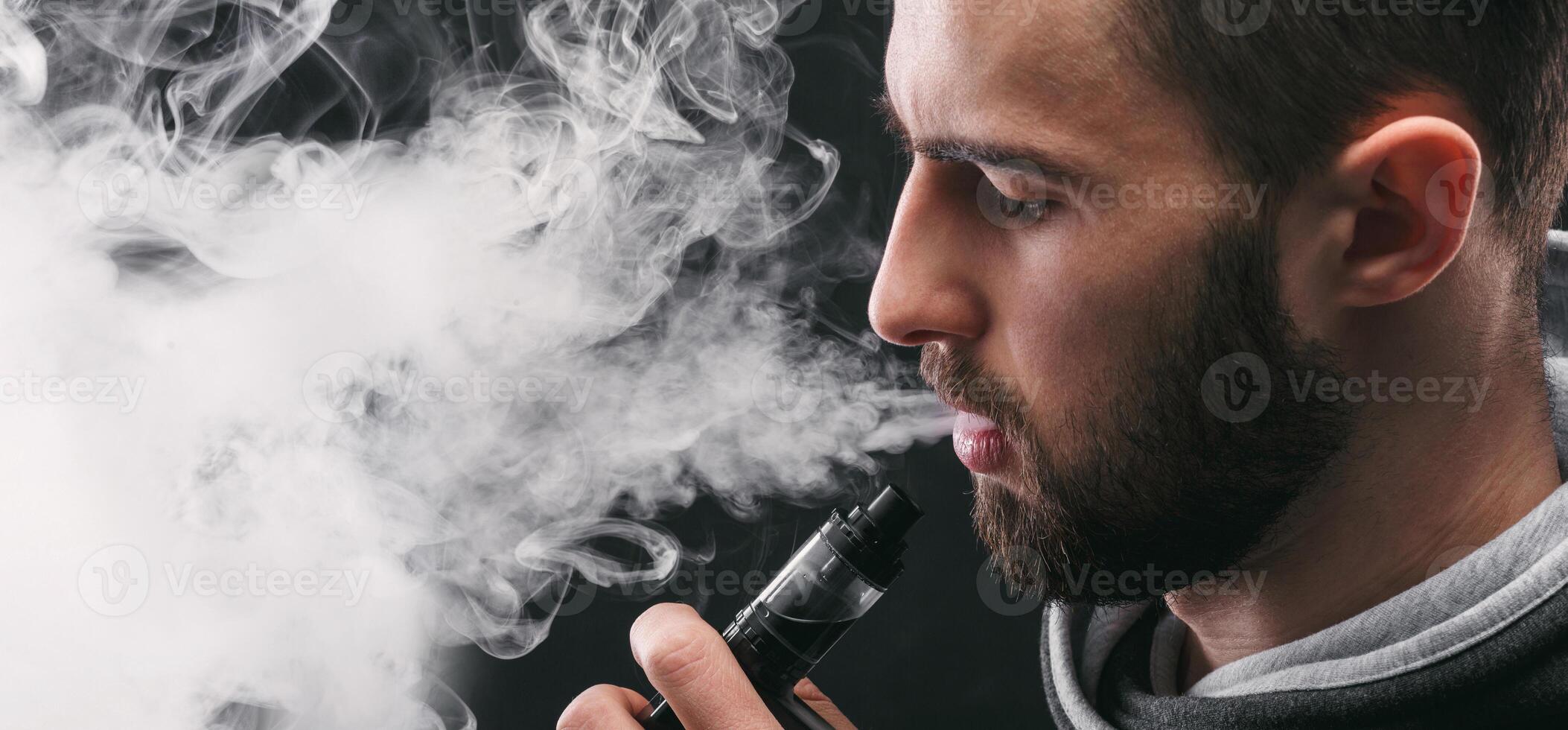 Man with vaping mod exhaling steam at black studio background. Bearded guy smoking e-cigarette to quit tobacco. Vapor and alternative nicotine free smoking concept, closeup photo
