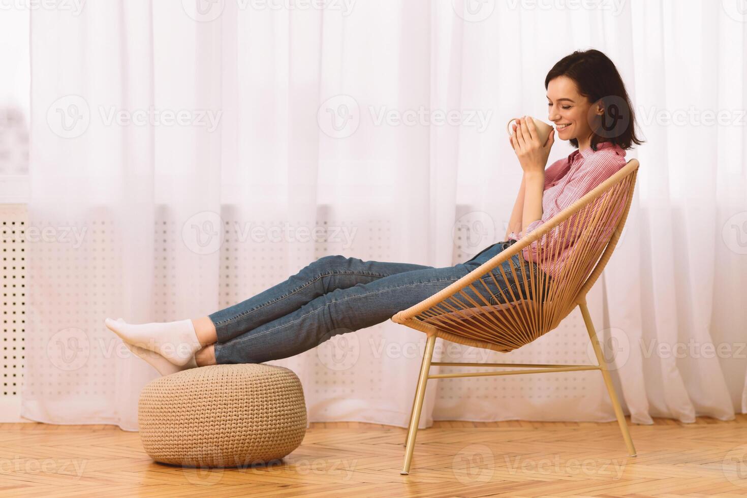 Weekend. Happy young woman sitting in modern chair, enjoying hot coffee in front of window, side view, copyspace photo